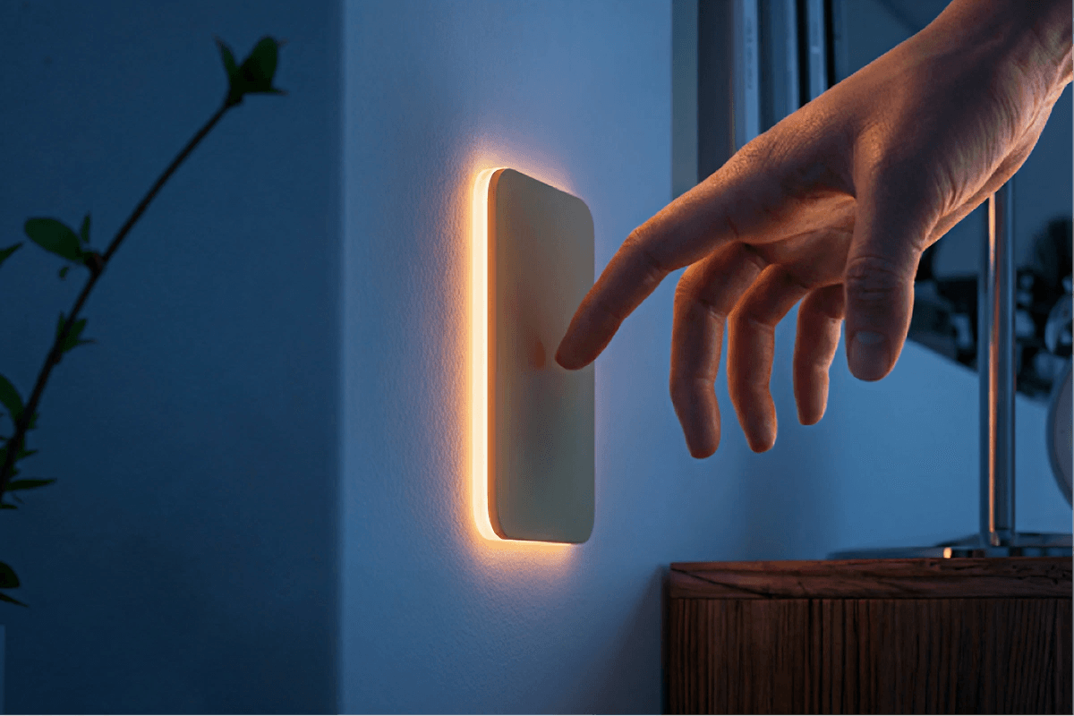 A human hand reaches out to touch a modern glowing smart light switch on a dark wall.