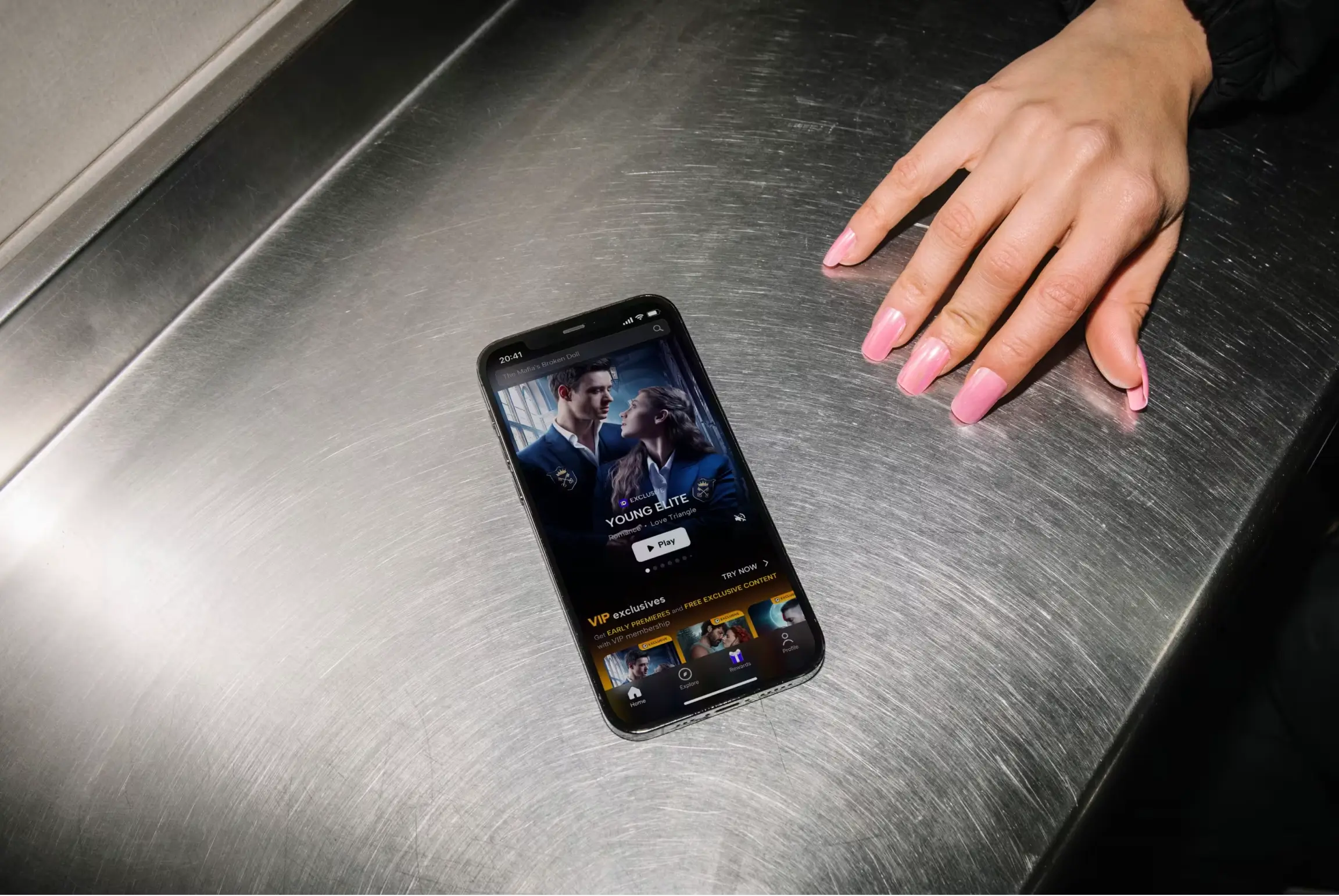Hand with pink nails next to a smartphone displaying a streaming app on a metallic surface.