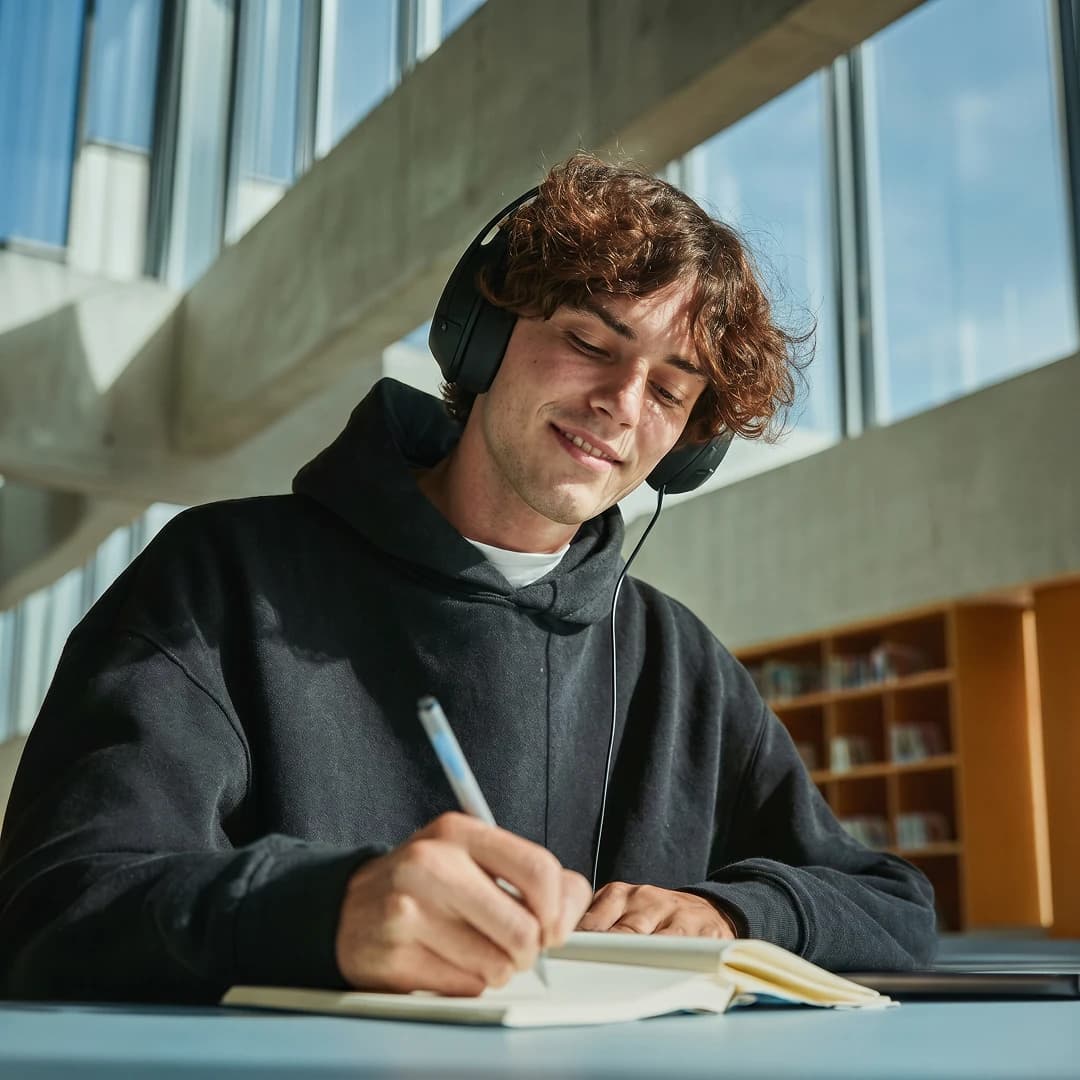Student with headphones writing in a notebook at a desk