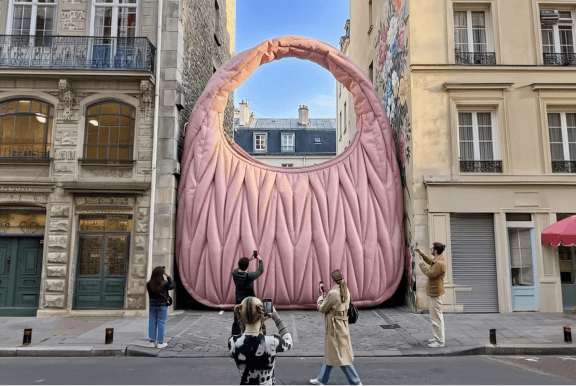 A massive pink quilted handbag sculpture stands between city buildings, with people photographing it.