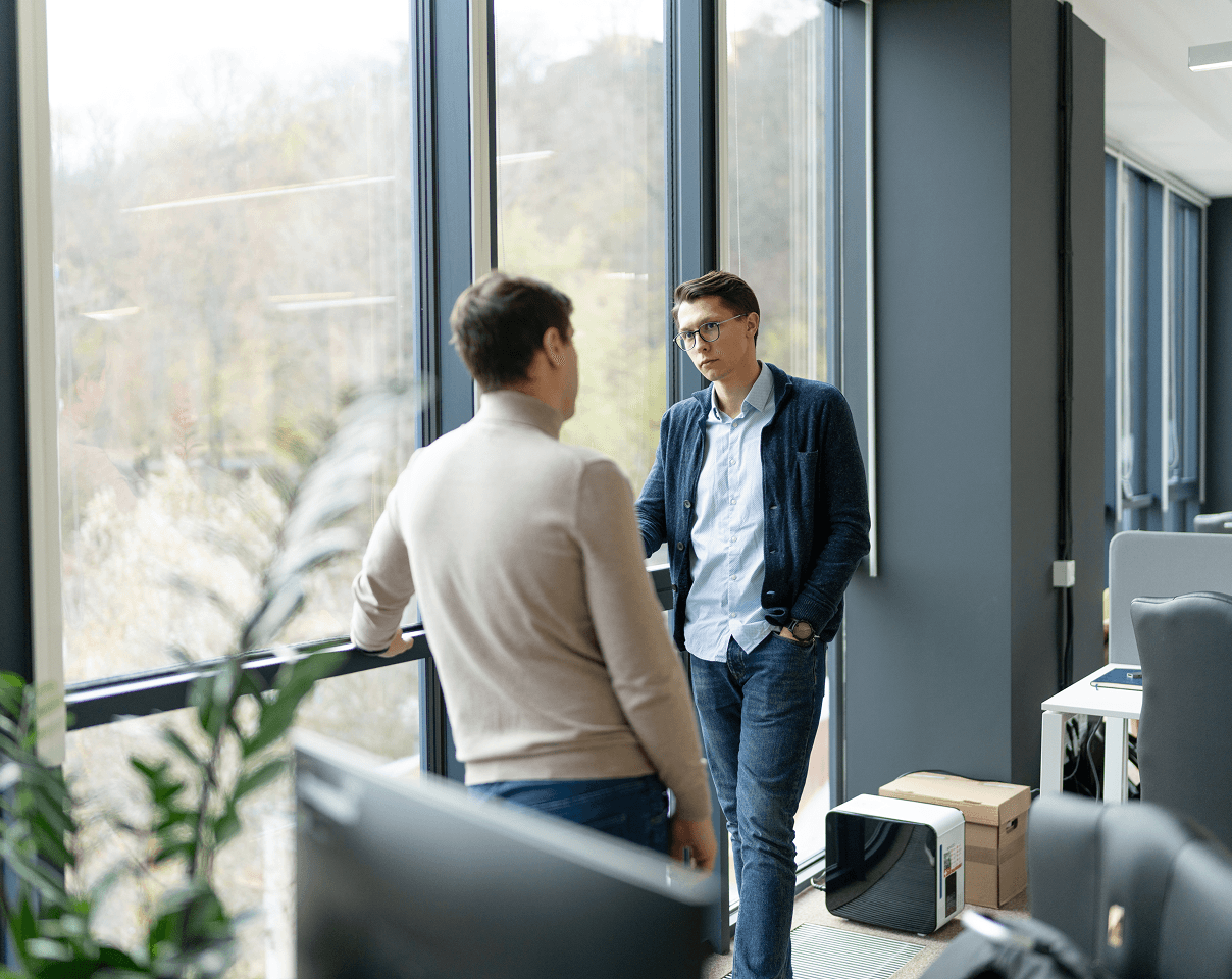 Two men in professional attire stand by a large window talking in an office.