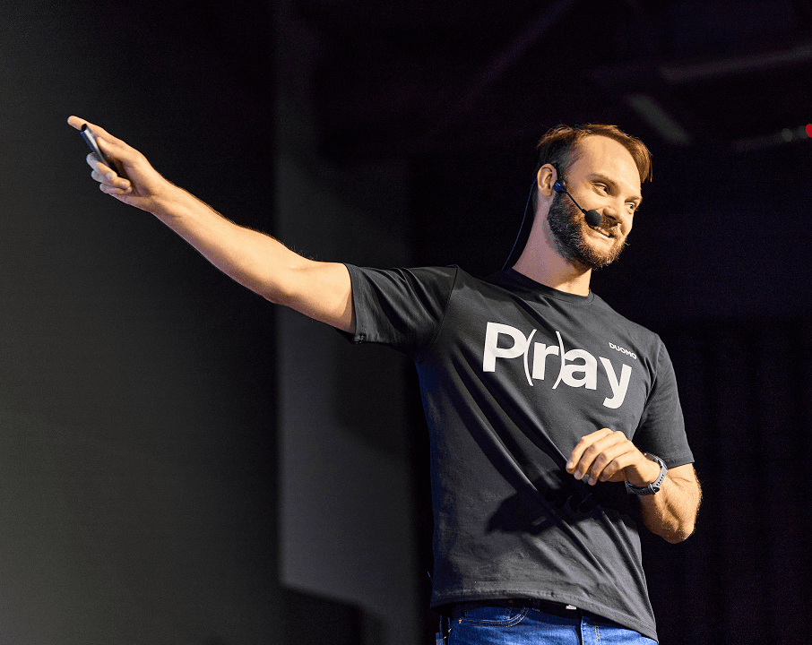 A male presenter on stage enthusiastically points to the side during a conference.