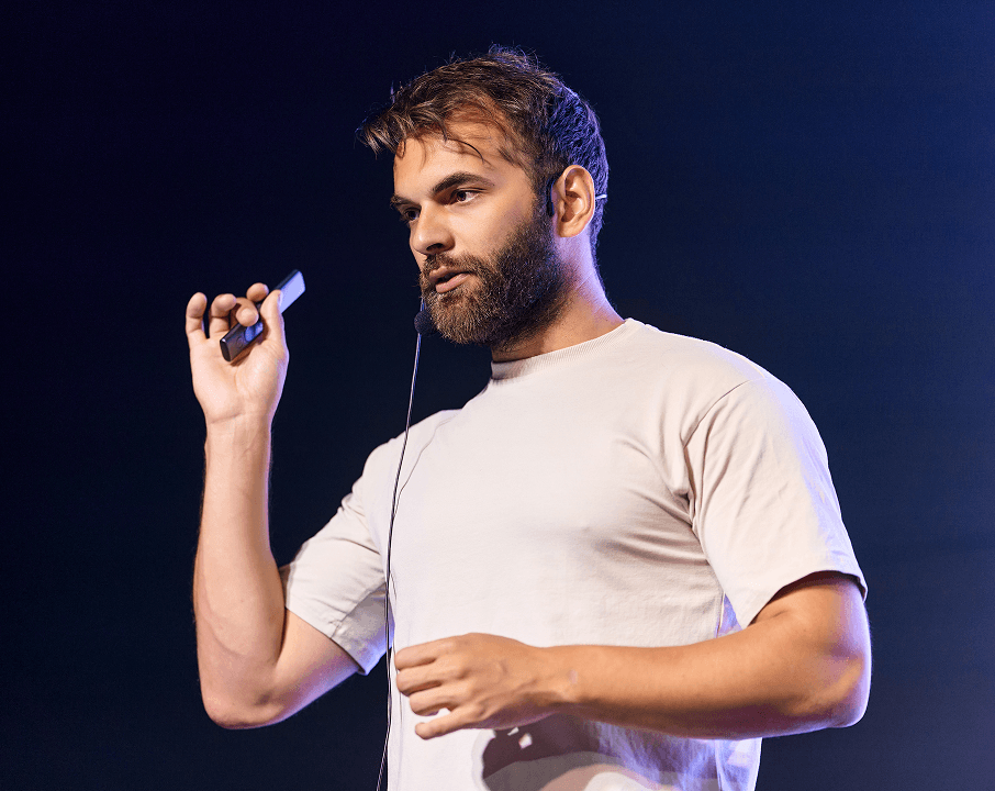A bearded man on stage holds a presentation clicker, looking engaged.