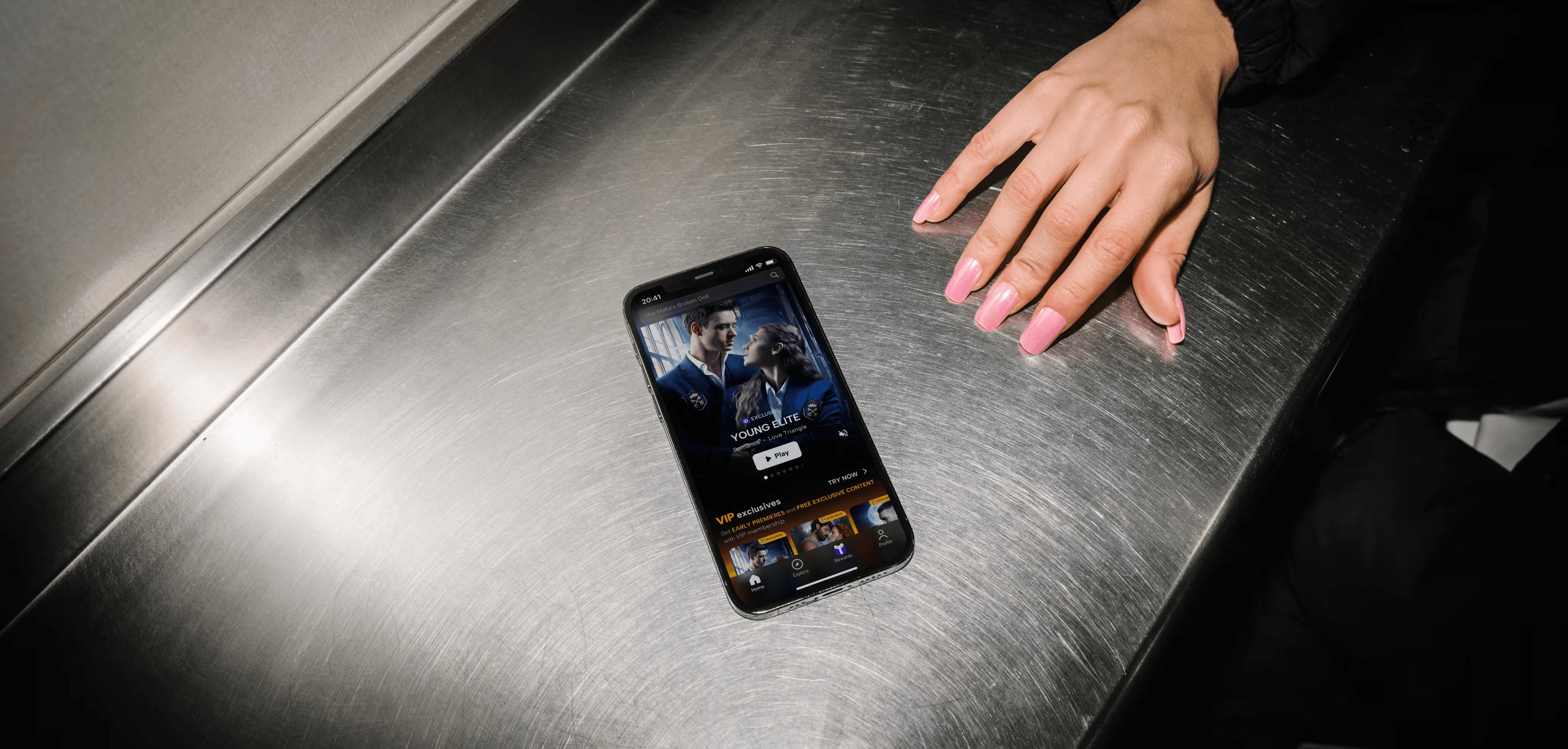 Smartphone on metal surface displaying streaming content, a hand with pink nails nearby.