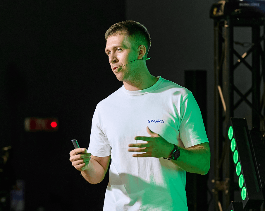 Man in white t-shirt speaking on stage with microphone, illuminated by green lights.