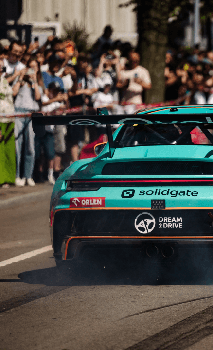 Teal and orange race car accelerating on a track, smoke from tires, crowd visible.
