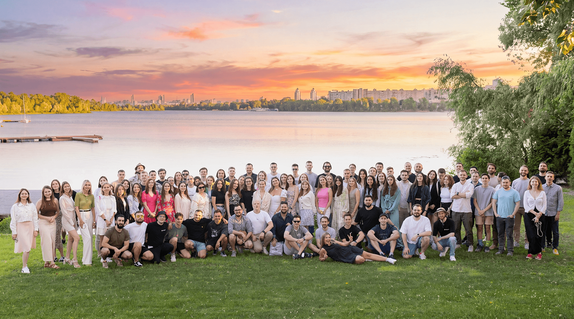 Large group of people posing for a photo on grass by a lake at sunset.